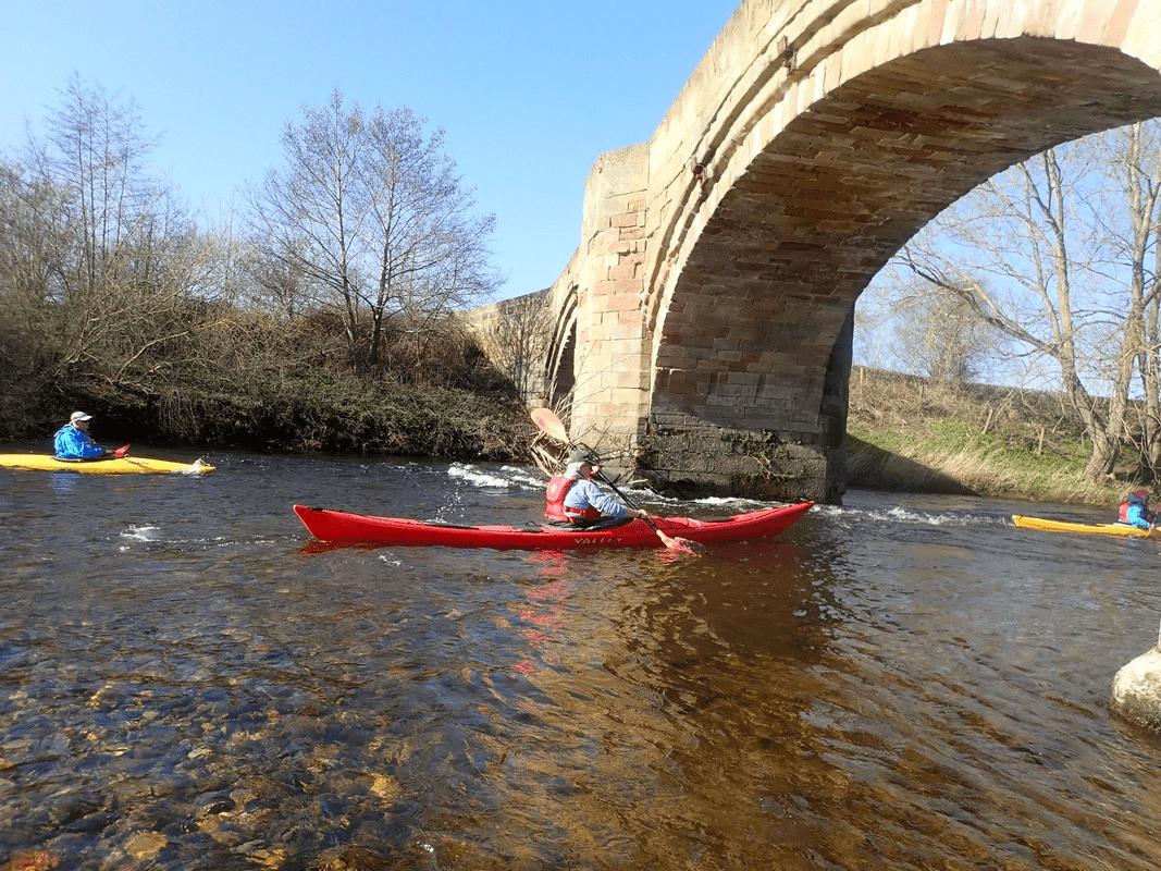 A group of people in kayaks on a river with a bridge in the backgroundDescription automatically generated with low confidence
