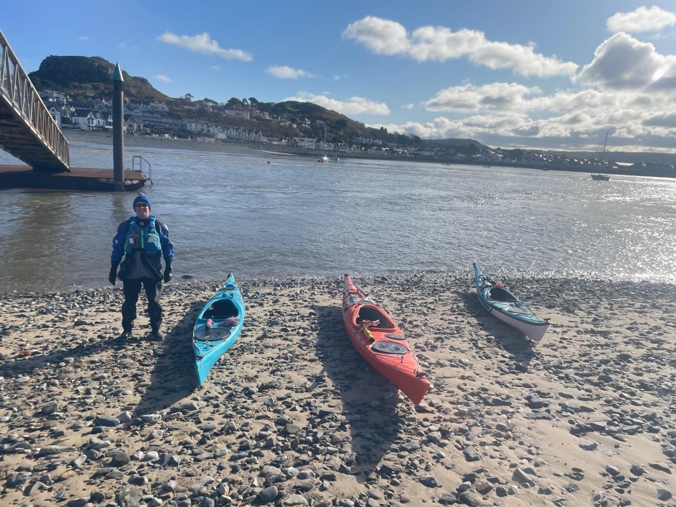 A person standing next to a row of kayaks on a beachDescription automatically generated with medium confidence