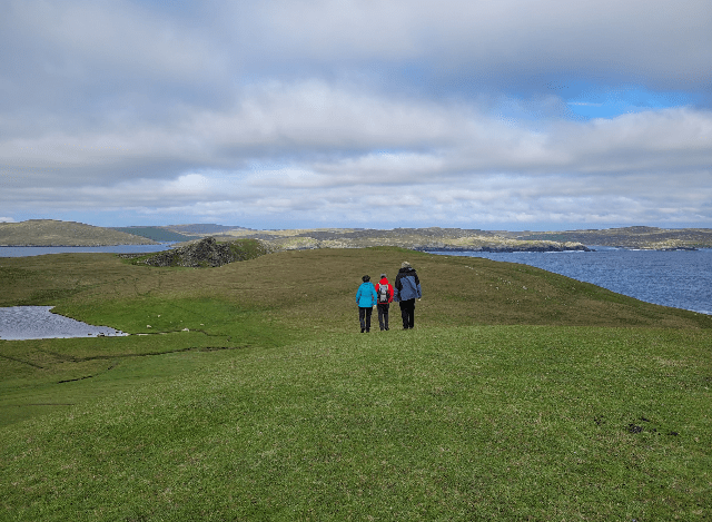 A group of people walking on a grassy hill by waterDescription automatically generated with medium confidence