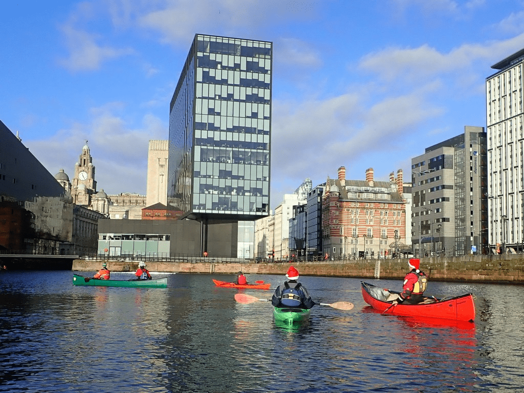 A group of people in kayaks in a river with buildings in the backgroundDescription automatically generated with low confidence