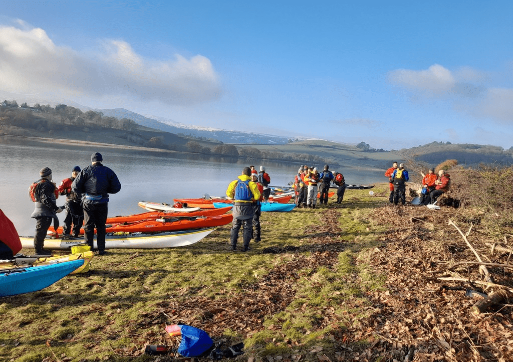 A group of people standing next to kayaks on a rocky shoreDescription automatically generated with low confidence