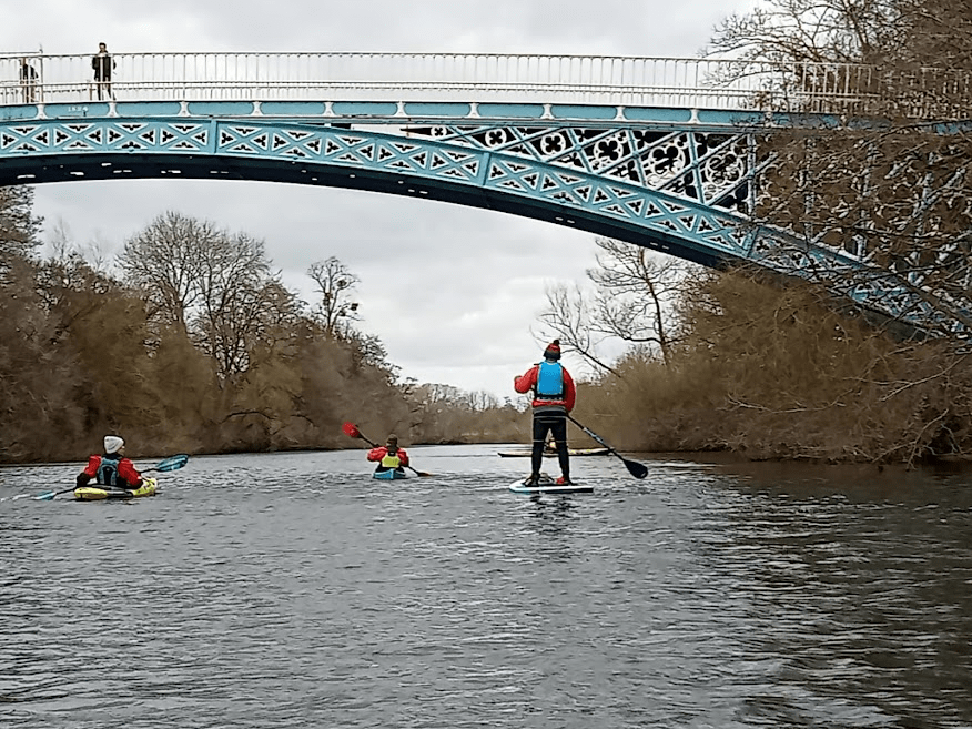A group of people paddle kayaks under a bridgeDescription automatically generated with medium confidence