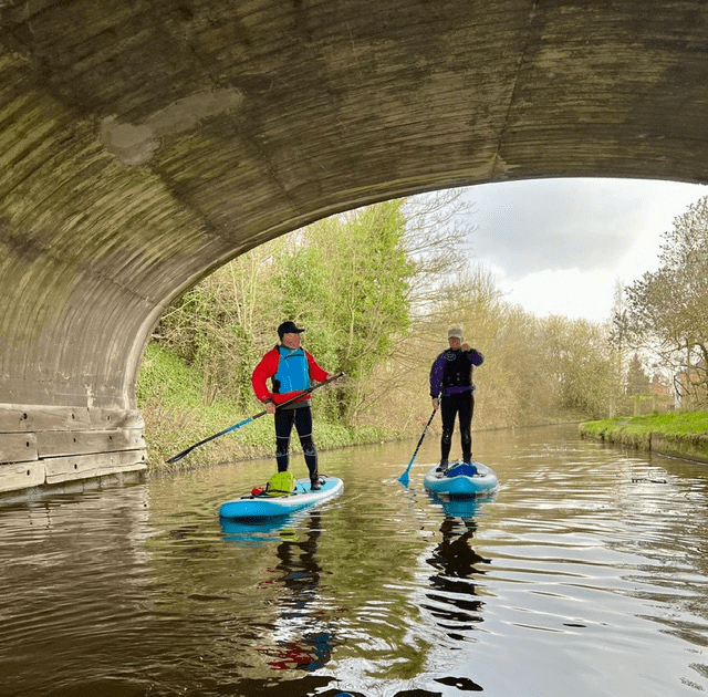 A couple of people on paddle boards in a river under a bridgeDescription automatically generated with low confidence