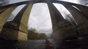 A person in a canoe under a bridge Description automatically generated