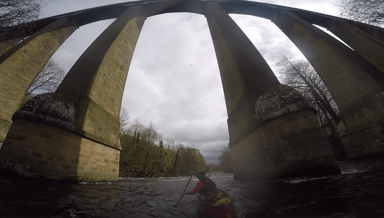 A person in a canoe under a bridge Description automatically generated