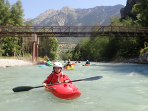 A group of people in kayaks on a river under a bridge Description automatically generated