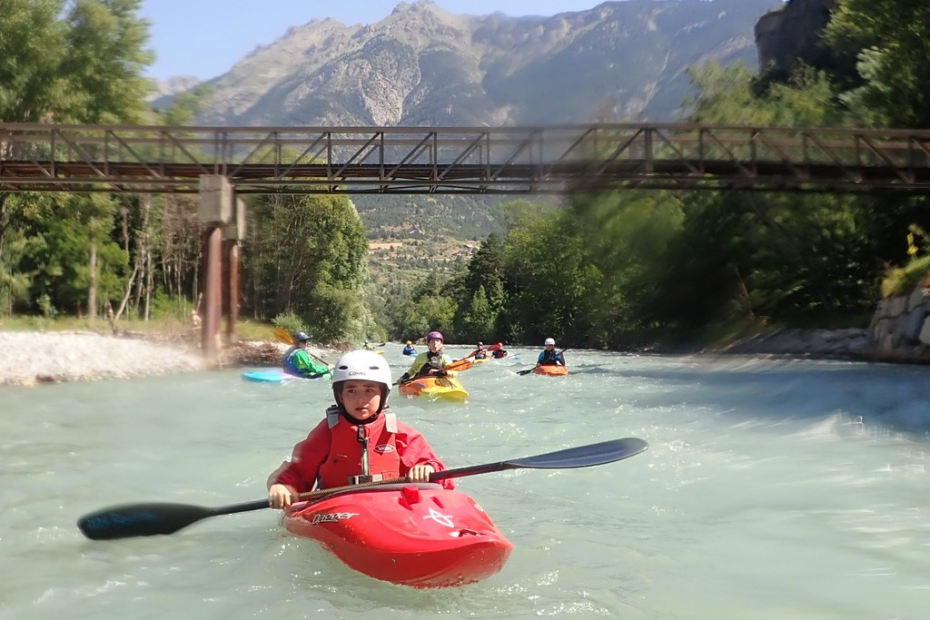 A group of people in kayaks on a river under a bridge Description automatically generated